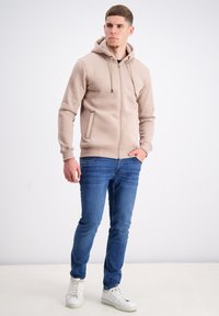 Šviesiai ruda zip-up hoodie su raišteliu, derinama su mėlynomis džinsinėmis kelnėmis ir baltais sportbačiais. Turi šonines kišenes ir glotnią tekstūrą.