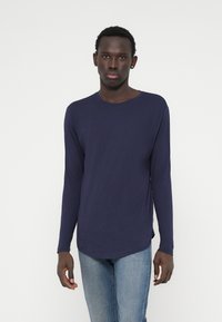 Marineblauw langemouwen shirt van zachte stof met een ronde hals, zijsplitten en een ontspannen pasvorm, gecombineerd met lichtblauwe jeans.