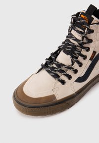 Beige høyhælte Vans-sko med svarte snøring, brun gummisåle, svart side stripe og oransje Vans-logo på siden og tungen.