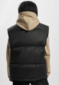 Schwarze Steppweste mit glänzender Textur, featuring einen hohen Kragen und ärmelloses Design, kombiniert mit einem beigen Hoodie.