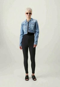 Cropped denim jacket with a light blue wash, featuring button detailing and two front pockets; paired with black leggings and black slide sandals.