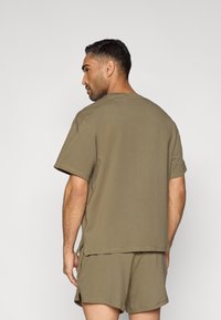 Πράσινο ελιάς βαμβακερό μπλουζάκι με κοντά μανίκια, στρογγυλή λαιμόκοψη και χαλαρή εφαρμογή, εμφανιζόμενο από πίσω, σε συνδυασμό με αντίστοιχα σορτς.