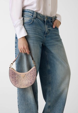 Personne portant un jean large bleu clair et une chemise blanche à manches longues, tenant un sac à main rose en forme de croissant orné de strass.