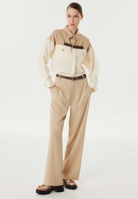 Giacca beige corta con maniche bianche, dotata di due tasche sul petto e una cintura di accento marrone. Abbinata a pantaloni beige larghi e sandali neri con zeppa.