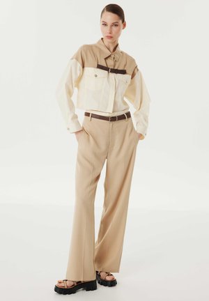 Giacca beige corta con maniche bianche, dotata di due tasche sul petto e una cintura di accento marrone. Abbinata a pantaloni beige larghi e sandali neri con zeppa.