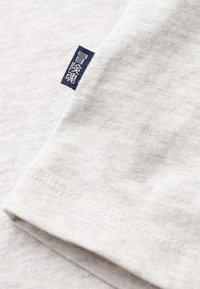 Lys grå bomulds t-shirt stof med fin struktur, med et marineblå label syet i kanten, der fremhæver detaljer og design.