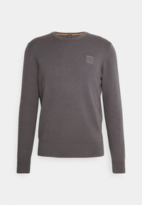 Grå stickad tröja med rundad halsringning, ribbade ärmslut och nederkant. Har en liten texturerad logopatch på övre vänstra sidan.