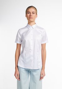 Chemise blanche à manches courtes avec un col pointu et une texture lisse et brillante. Elle présente une coupe ajustée et un plastron à cinq boutons.