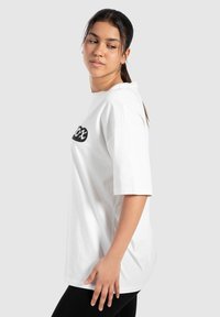 Camiseta de algodón blanca con un logo negro, de corte relajado, con mangas cortas y un escote redondo, exhibida sobre un fondo neutro.