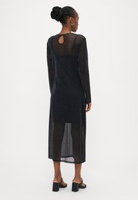 Μαύρο φόρεμα με μακριά μανίκια από διάφανο, λαμπερό ύφασμα. Διαθέτει άνοιγμα σε σχήμα κλειδιού στην πλάτη και ίσια γραμμή. Φορεμένο με μαύρα σανδάλια με χοντρό τακούνι.