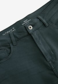 Pantaloni strâmți de culoare verde închis, cu o textură fină, închidere cu buton metalic și detalii cusute, având branding pe talie.
