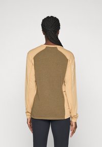 Persoon met gevlochten haar in een lange beige en olijfgroene top met "Peak Performance" op de rechtermouw, met de rug naar de camera gekeerd.