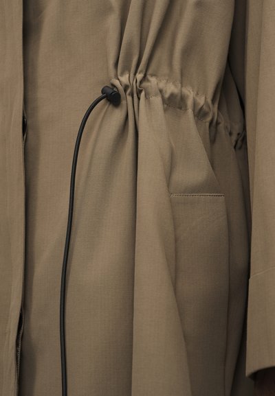 Khaki tkanina sťahovaná šnúrkou a detailom vrecka na odeve, zdôrazňujúca textúru a šitie.