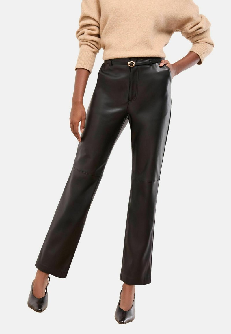 Sieviete valkā melnas augstās imitācijas ādas bikses ar jostu, beigu krāsas džemperi ar uzripinātām piedurknēm un melnas špikotu augstpapēžu kurpes.