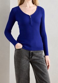 Haut bleu à manches longues côtelé avec un col en V et une fermeture éclair frontale. Tissu texturé ; associé à un jean gris à coupe décontractée.