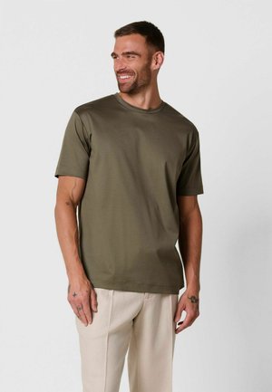 Uomo che sorride e guarda a sinistra indossando una maglietta verde oliva a maniche corte e pantaloni beige chiaro su sfondo semplice.