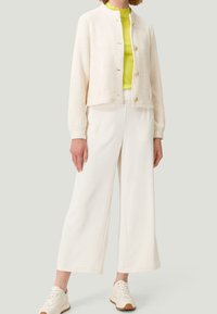 Giacca lavorata a maglia color crema con bottoni dorati, indossata sopra una maglia giallo brillante e pantaloni bianchi a gamba larga, abbinata a sneakers chiare.