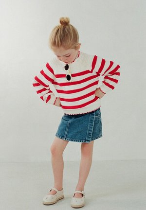 Jeune fille aux cheveux blonds attachés en chignon, portant un pull rayé rouge et blanc, une jupe en jean, des chaussures blanches et des lunettes de soleil accrochées à sa poitrine, regardant vers le bas.