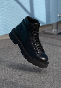 Botte en cuir noir à lacets avec une épaisse semelle en caoutchouc suspendue au-dessus d'un sol en asphalte, près d'un mur texturé bleu clair.