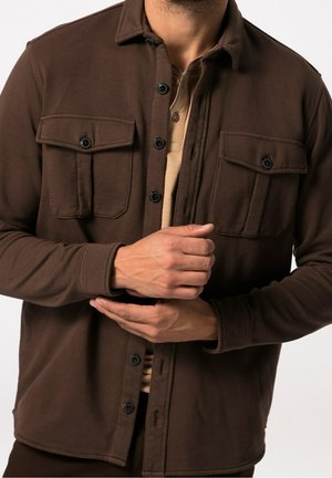 Mann trägt eine braune Knopfleistenjacke mit vorderen Klappentaschen und ein beiges Hemd, die Hände vor der Taille verschränkt, vor einfachem Hintergrund.