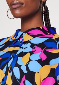 Färggshirt med blommönster i blå, rosa och orangea blad mot en svart bakgrund; innehåller krage och knappdetaljer.