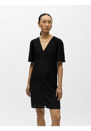 Femme debout portant une robe noire texturée longueur genou avec des manches courtes, un col en V, des boutons à l'avant, des boucles d'oreilles créoles et un bracelet en argent.