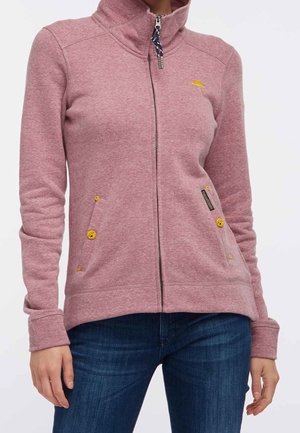 Mujer con chaqueta de forro polar malva con cremallera y detalles de botones amarillos y vaqueros azules, de pie frente a un fondo liso.
