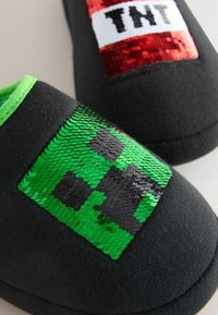 Black slippers featuring green and black sequin designs with a pixelated pattern, accented by a red and white "TNT" patch. Green interior lining.