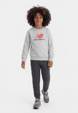 Görög hajú gyermek szürke New Balance pulóvert, sötétszürke melegítőnadrágot és piros részletekkel díszített szürke sportcipőt viselve sétál előre.