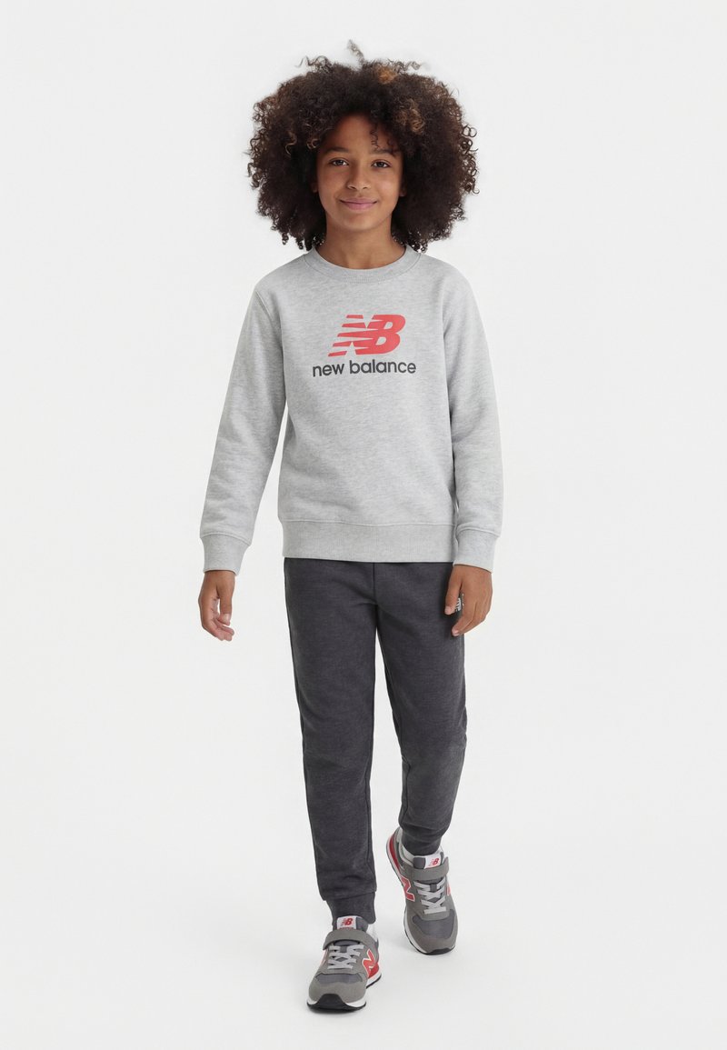 Παιδί με κατσαρά μαλλιά που περπατάει προς τα εμπρός, φορώντας γκρι φούτερ New Balance, σκούρες γκρι φόρμες και γκρι αθλητικά παπούτσια με κόκκινες λεπτομέρειες.