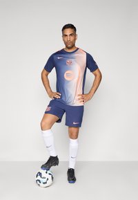 Marinblå fotballdrakt med gradient oransje detaljer, rund hals og logo. Svarte fotballsko og hvite strømper. En fotball ligger på bakken.