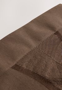 Közelkép barna bordás szövetről, melyen texturált halszálkaminta található finom párhuzamos vonalakkal és változatos kötött öltésekkel.