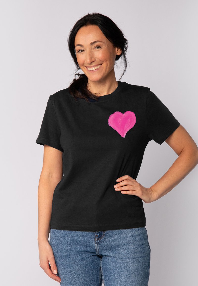 Zwarte katoenen t-shirt met een groot roze hartontwerp op de linker borstzijde, ronde halslijn en korte mouwen. Model lacht.