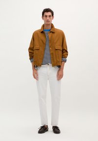 Chaqueta de algodón marrón con dos grandes bolsillos, superpuesta a una camisa de rayas azul y blanca, combinada con jeans blancos y zapatos oscuros.