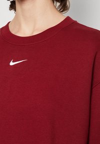Bordowy sweter z okrągłym dekoltem i prążkowanym wykończeniem. Posiada małe, białe logo Nike po lewej stronie. Miękka tekstura materiału i luźny krój.