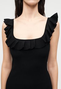 Γυναίκα φοράει μαύρο αμάνικο τοπ με φαρδιά τετράγωνη λαιμόκοψη και βολάν κατά μήκος της λαιμόκοψης και των ώμων.