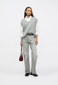 Γκρι πλεκτό αμάνικο πουλόβερ πάνω από ένα λευκό πουκάμισο, συνδυασμένο με ανοιχτού γκρι παντελόνι και μαύρα παπούτσια. Κρατά μια μικρή μπορντό τσάντα.