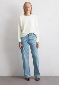 Licht crèmekleurige trui met lange mouwen, gecombineerd met lichtblauwe rechte jeans. De outfit heeft een ontspannen pasvorm en wordt aangevuld met zwarte schuifslippers.