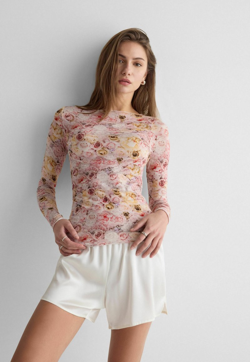 Junge Frau trägt ein langärmliges, florales Oberteil und weiße Shorts, posiert vor einem schlichten, hellgrauen Hintergrund.