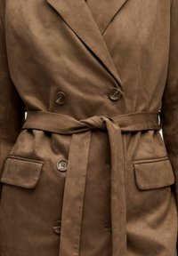 Manteau double boutonnage en daim marron avec quatre boutons, grands revers, poches à rabat et une ceinture en tissu nouée à la taille.