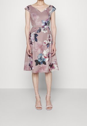 Femme portant une robe fleurie mauve longueur genou avec manches courtes et encolure en V, associée à des escarpins rose pâle à bride à la cheville, debout contre un mur blanc.