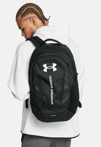 Sac à dos noir Under Armour avec une surface texturée, arborant un logo blanc, des bretelles réglables et une poche latérale en filet pour le rangement.
