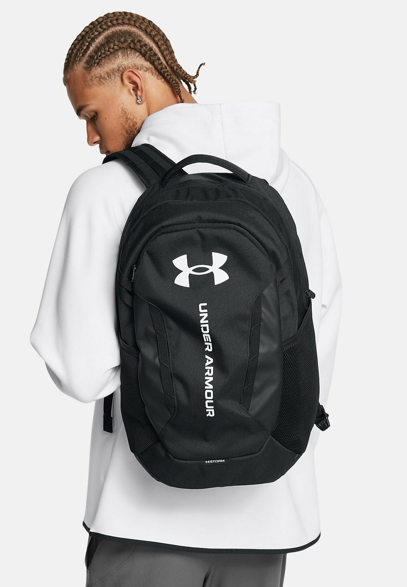 Sac à dos noir Under Armour avec une surface texturée, arborant un logo blanc, des bretelles réglables et une poche latérale en filet pour le rangement.