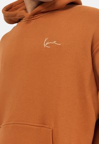 Felpa arancione in cotone, con tasca frontale e logo ricamato bianco sul petto. Tessuto morbido con design casual.