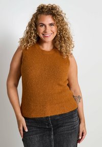 Femme souriante avec des cheveux blonds bouclés portant un débardeur en tricot marron sans manches et un jean foncé, montrant un tatouage de rose sur l'avant-bras gauche.