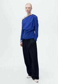 Top blu off-the-shoulder con design drappeggiato, abbinato a pantaloni neri larghi. Texture liscia, stile minimalista e sottili accenti ai polsi.