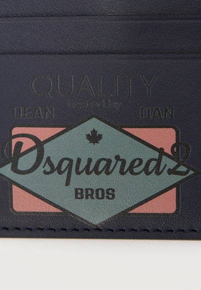Σκούρο μπλε δερμάτινη ετικέτα με την λέξη "QUALITY" ανάγλυφη πάνω από ένα διαμάντι που περιέχει τις λέξεις "Dsquared2 BROS" και ένα μικρό έμβλημα με φύλλο σφενδάμου.