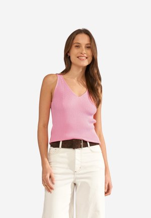 Femme souriante, portant un débardeur rose côtelé à col en V, un pantalon blanc taille haute et une ceinture marron foncé, avec des cheveux bruns lâchés.