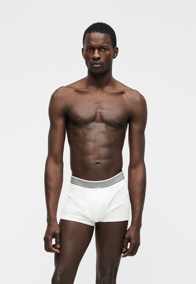 Homme musclé à la peau foncée, debout face à l'avant, portant un boxer blanc avec une ceinture décorative, sur un fond clair uni.