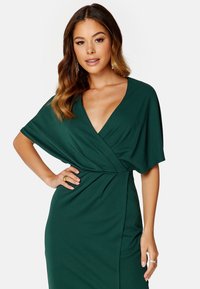 Groene wikkeljurk met korte vleermuismouwen, diepe V-hals en een aansluitende taille. Zachte, gladde stof met subtiele draperingdetails.